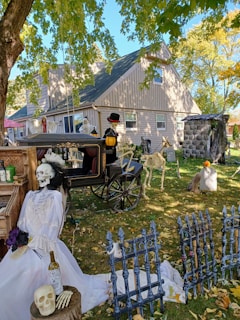A Halloween-themed scene with skeletons dressed in costumes, a horse-drawn carriage decorated with a skeleton driver, and various props like a skull, bottle, and tombstones in a yard. The setting is a suburban house under the shade of trees with fallen autumn leaves.