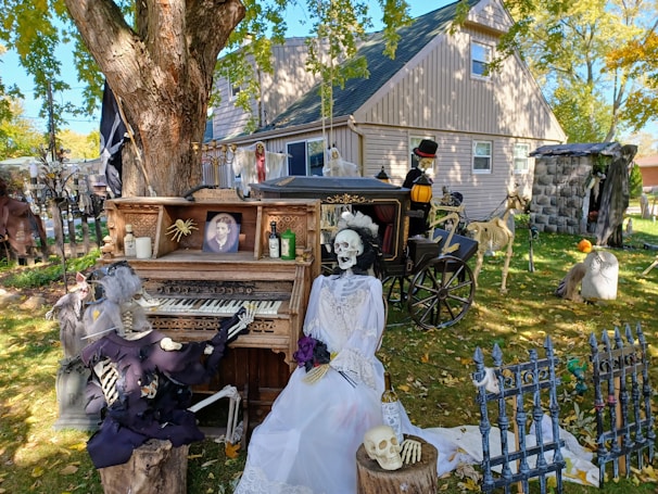 The image depicts a Halloween-themed outdoor scene in front of a house. There is a large tree with a detailed wooden piano beneath it, on which a skeleton is playing. Another skeleton dressed as a bride is seated on a bench beside the piano, holding a purple bouquet. The scene also includes various other Halloween decorations like skulls, bottles, a fence, and several costumes. A horse-drawn carriage with a driver skeleton is visible in the background, and the yard is littered with other spooky figurines and props.