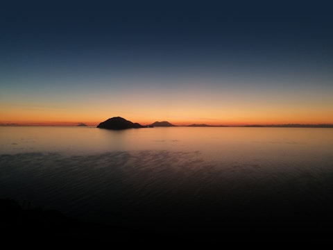 The image features a breathtaking sunset over a calm body of water, with a dark silhouette of a distant island and other landforms on the horizon. The sky transitions from deep blue at the top to vibrant orange and yellow near the horizon, casting a warm glow.