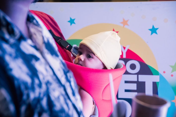 A baby is seen wearing a yellow hat and looking curiously from a bright red baby carrier. The background features a colorful sign with stars and large text.