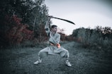 A person is wearing traditional martial arts attire and is executing a wide stance while wielding a large sword above their head. The background features an outdoor scene with dense foliage displaying green and deep red hues, indicative of a forested or wooded area.