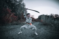 A person is wearing traditional martial arts attire and is executing a wide stance while wielding a large sword above their head. The background features an outdoor scene with dense foliage displaying green and deep red hues, indicative of a forested or wooded area.
