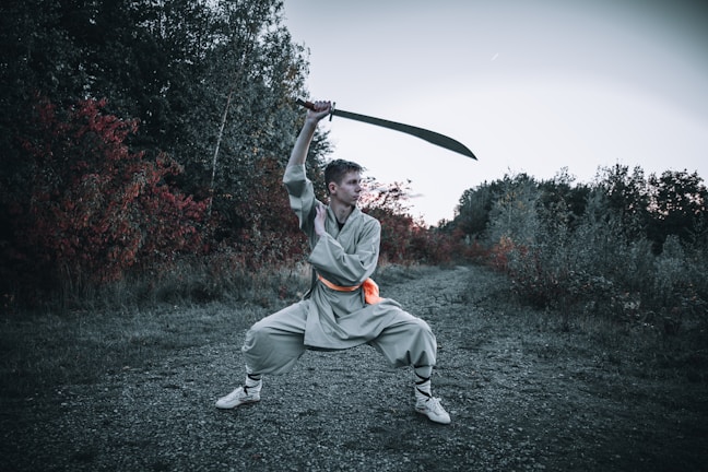 A person is wearing traditional martial arts attire and is executing a wide stance while wielding a large sword above their head. The background features an outdoor scene with dense foliage displaying green and deep red hues, indicative of a forested or wooded area.