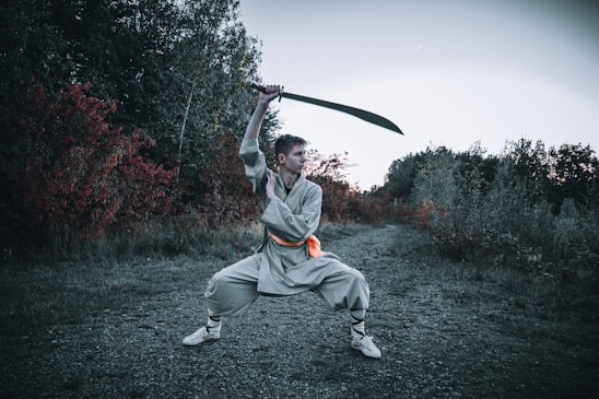 A person is wearing traditional martial arts attire and is executing a wide stance while wielding a large sword above their head. The background features an outdoor scene with dense foliage displaying green and deep red hues, indicative of a forested or wooded area.