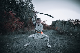 A person is wearing traditional martial arts attire and is executing a wide stance while wielding a large sword above their head. The background features an outdoor scene with dense foliage displaying green and deep red hues, indicative of a forested or wooded area.