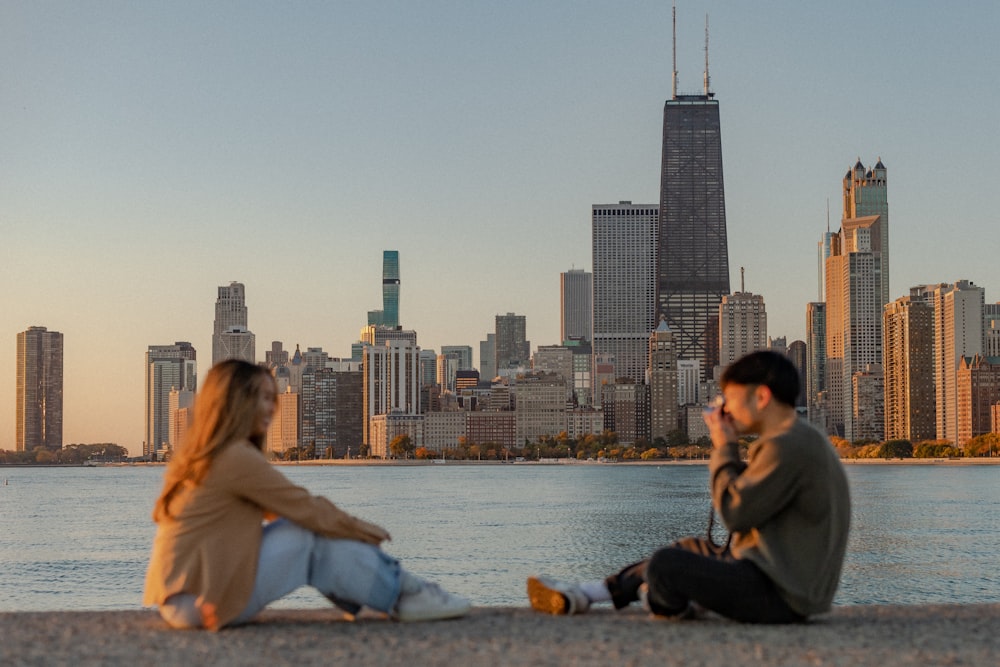 a man and woman sitting on a beach with a city skyline in the background