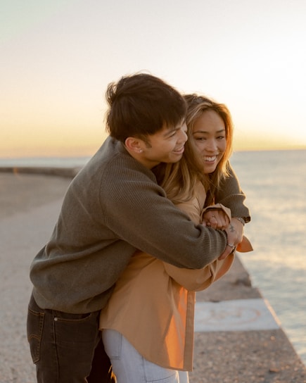 A young couple embraces happily by a seaside walkway during a sunset. The background shows calm water and a clear sky, creating a warm and romantic atmosphere.