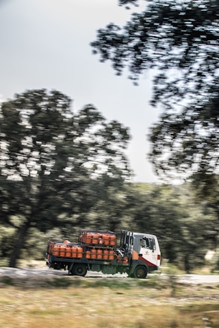 A truck is carrying several gas cylinders, driving along a road bordered by trees. The background is slightly blurred, indicating motion.