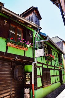 A quaint, half-timbered house features a vibrant green facade with wooden frames and a clay-tiled roof. Bright flowers in window boxes add color, and wooden barrels and sculptures are visible outside. A traditional signboard displays text in French.