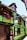 A quaint, half-timbered house features a vibrant green facade with wooden frames and a clay-tiled roof. Bright flowers in window boxes add color, and wooden barrels and sculptures are visible outside. A traditional signboard displays text in French.