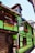 A quaint, half-timbered house features a vibrant green facade with wooden frames and a clay-tiled roof. Bright flowers in window boxes add color, and wooden barrels and sculptures are visible outside. A traditional signboard displays text in French.