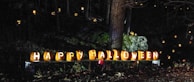Carved pumpkins with illuminated letters spelling 'Happy Halloween' are arranged in a row. They are surrounded by additional jack-o'-lanterns scattered through a dimly lit forest setting, with trees and rocks in the background.