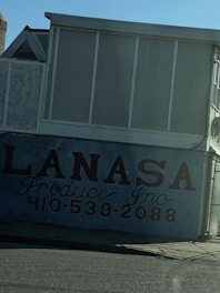 A building facade featuring a large sign with the text 'LANASA Produce Inc.' and a phone number beneath it. The structure is a combination of dark and light shades, with a slanted sidewalk in front. The overall scene is under a clear blue sky with no visible people.