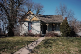 A quaint, rustic house with a peaked roof and decorative trim sits in a rural setting. The home has a small porch with wooden railings and is surrounded by bare trees and shrubs. The sky is clear, enhancing the peaceful ambiance of the yard, which features a narrow concrete pathway leading to the entrance.