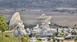 A large complex of satellite dishes of varying sizes is situated in an open field surrounded by hills. The main structure features multiple large parabolic antennas, suggesting a communication or space observation facility. The setting is rural with sparse vegetation and some small roads leading to the complex.