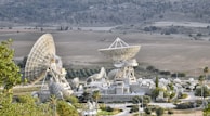 A large complex of satellite dishes of varying sizes is situated in an open field surrounded by hills. The main structure features multiple large parabolic antennas, suggesting a communication or space observation facility. The setting is rural with sparse vegetation and some small roads leading to the complex.