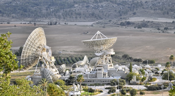 A large complex of satellite dishes of varying sizes is situated in an open field surrounded by hills. The main structure features multiple large parabolic antennas, suggesting a communication or space observation facility. The setting is rural with sparse vegetation and some small roads leading to the complex.