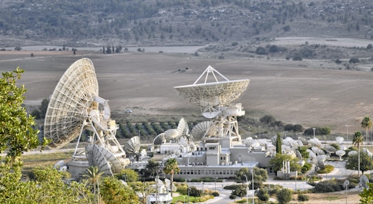 A large complex of satellite dishes of varying sizes is situated in an open field surrounded by hills. The main structure features multiple large parabolic antennas, suggesting a communication or space observation facility. The setting is rural with sparse vegetation and some small roads leading to the complex.