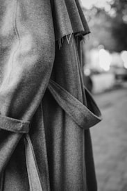 A close-up of a textured coat with frayed edges and a belt, captured in black and white. The background is softly blurred, highlighting the fabric's intricate details and the garment's stylish design.