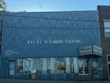 A brick building with a large sign reading 'WRF & S N Plumbing & Heating' on the front facade. There are advertisements for Goulds Pumps, water conditioners, and supplies on the wall. The storefront features large glass windows displaying plumbing-related items. The door on the left is labeled '15 N Main' and there is a sign indicating propane cylinder refills.