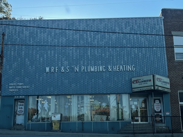 A brick building with a large sign reading 'WRF & S N Plumbing & Heating' on the front facade. There are advertisements for Goulds Pumps, water conditioners, and supplies on the wall. The storefront features large glass windows displaying plumbing-related items. The door on the left is labeled '15 N Main' and there is a sign indicating propane cylinder refills.