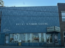 A brick building with a large sign reading 'WRF & S N Plumbing & Heating' on the front facade. There are advertisements for Goulds Pumps, water conditioners, and supplies on the wall. The storefront features large glass windows displaying plumbing-related items. The door on the left is labeled '15 N Main' and there is a sign indicating propane cylinder refills.