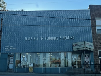 A brick building with a large sign reading 'WRF & S N Plumbing & Heating' on the front facade. There are advertisements for Goulds Pumps, water conditioners, and supplies on the wall. The storefront features large glass windows displaying plumbing-related items. The door on the left is labeled '15 N Main' and there is a sign indicating propane cylinder refills.
