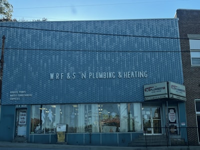 A brick building with a large sign reading 'WRF & S N Plumbing & Heating' on the front facade. There are advertisements for Goulds Pumps, water conditioners, and supplies on the wall. The storefront features large glass windows displaying plumbing-related items. The door on the left is labeled '15 N Main' and there is a sign indicating propane cylinder refills.