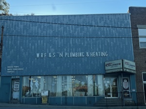 A brick building with a large sign reading 'WRF & S N Plumbing & Heating' on the front facade. There are advertisements for Goulds Pumps, water conditioners, and supplies on the wall. The storefront features large glass windows displaying plumbing-related items. The door on the left is labeled '15 N Main' and there is a sign indicating propane cylinder refills.