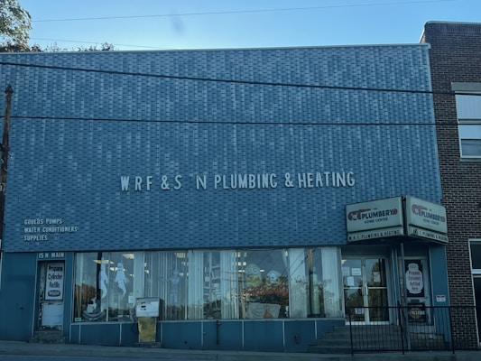 A brick building with a large sign reading 'WRF & S N Plumbing & Heating' on the front facade. There are advertisements for Goulds Pumps, water conditioners, and supplies on the wall. The storefront features large glass windows displaying plumbing-related items. The door on the left is labeled '15 N Main' and there is a sign indicating propane cylinder refills.