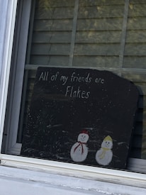 A rectangular blackboard is displayed in a window with horizontal blinds in the background. The blackboard has a humorous message written in white chalk, stating 'All of my friends are flakes,' accompanied by drawings of two snowmen, one with a red scarf and cap and the other with a yellow scarf and cap.