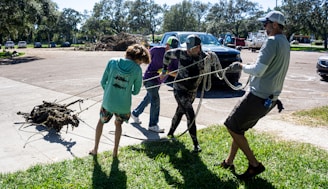 A group of people are engaged in an outdoor activity, pulling a heavy object with ropes. They are standing on a grass and concrete surface in a parking lot. Several vehicles are parked in the background, and trees surround the area.