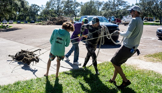 A group of people are engaged in an outdoor activity, pulling a heavy object with ropes. They are standing on a grass and concrete surface in a parking lot. Several vehicles are parked in the background, and trees surround the area.