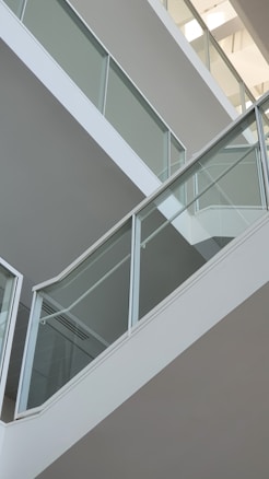 A modern architectural interior featuring a series of sleek, angled staircases with glass railings and white structural elements. Sunlight filters through, creating gentle shadows on the walls, adding depth to the minimalist design.