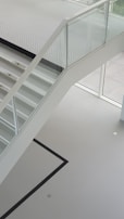 A modern staircase with glass railing and white steps is seen. The staircase has a minimalist design and is positioned in a bright, open space with large windows that allow natural light to enhance the clean lines. The floor features a simple, geometric pattern with a black line on a light surface.