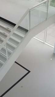 A modern staircase with glass railing and white steps is seen. The staircase has a minimalist design and is positioned in a bright, open space with large windows that allow natural light to enhance the clean lines. The floor features a simple, geometric pattern with a black line on a light surface.