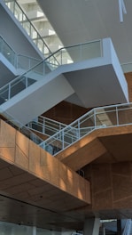 Modern architectural interior featuring a series of angular staircases with glass railings. The space is characterized by its geometric design and use of natural light, highlighting the contrast between white and wooden textures.