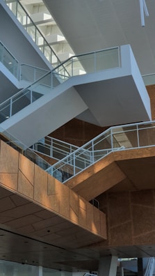 Modern architectural interior featuring a series of angular staircases with glass railings. The space is characterized by its geometric design and use of natural light, highlighting the contrast between white and wooden textures.