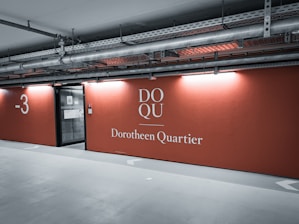 A parking garage area with concrete floors and ceilings, featuring industrial pipes and lighting fixtures above. The walls are painted orange, displaying the words 'DOQU Dorotheen Quartier' and the level '-3'. There is an entrance to an elevator or stairwell with a glass door on the left.