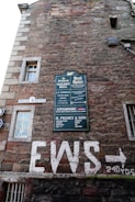 An exterior wall of an old brick building features multiple signs, including a large green sign with various business names, a white street sign reading High Wiend, and graffiti-like white letters spelling out EWS with an arrow pointing right. Windows with white frames are scattered on the wall, and there are multiple textures and colors in the masonry.