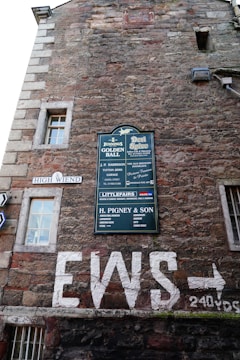 An exterior wall of an old brick building features multiple signs, including a large green sign with various business names, a white street sign reading High Wiend, and graffiti-like white letters spelling out EWS with an arrow pointing right. Windows with white frames are scattered on the wall, and there are multiple textures and colors in the masonry.