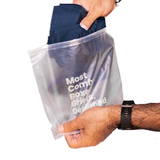 A hand wearing a wristwatch is holding a translucent plastic bag containing dark-colored clothing. The bag has text printed on it in white, advertising comfort and design.