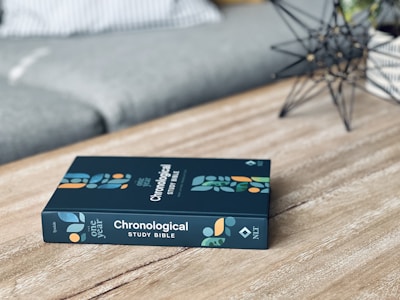 A book titled 'Chronological Study Bible' rests on a wooden table. The cover features geometric patterns in shades of orange, blue, and green. Nearby, a wireframe ornamental structure adds a decorative element to the scene, and a striped cushion is partially visible in the background.