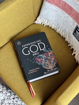 A book titled 'Experiencing God Bible' rests on a mustard yellow armchair. The book cover features an abstract colorful design resembling fire. Next to the book, a cozy gray and red striped blanket with tassels is draped over the arm of the chair.