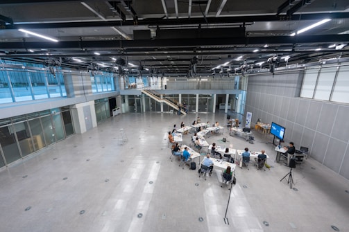 A large, modern conference room with high ceilings and an industrial look is depicted. There is a group of people sitting around rectangular tables arranged in a circular formation in the center of the room. They appear to be engaged in a meeting or workshop. The room has glass walls on the left side, and a staircase leading to an upper level. Several large screens and audio-visual equipment are set up, suggesting a formal presentation setting. The space is well-lit, with light fixtures visible in the ceiling.