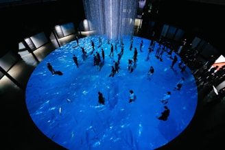 A large circular area with a blue surface resembling water, featuring many people walking around and interacting. In the center, hanging strands or light elements create a focal point. Surrounding the circle are dark walls with illuminated panels.
