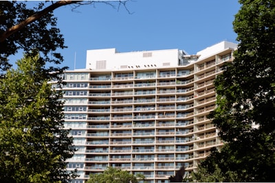 Modern glass-fronted apartment building with balconies surrounded by greenery under a clear blue sky