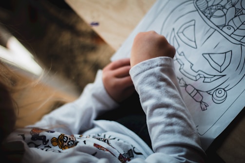 A child is coloring a Halloween-themed drawing depicting a jack-o'-lantern with a variety of smaller illustrations around it. They are holding a crayon or colored pencil and wearing a long-sleeved shirt with a printed design on it.