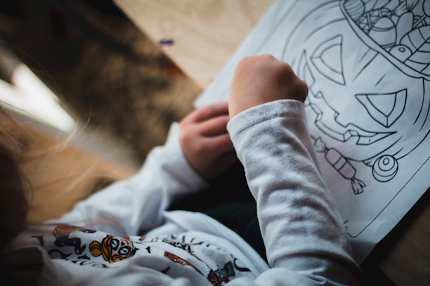 A child is coloring a Halloween-themed drawing depicting a jack-o'-lantern with a variety of smaller illustrations around it. They are holding a crayon or colored pencil and wearing a long-sleeved shirt with a printed design on it.