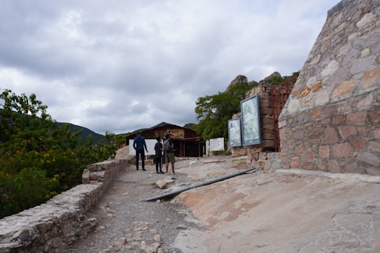 A rustic pathway leads to a wooden building with large signs displayed outside. Three people are walking toward the building, which is surrounded by lush greenery and mountains in the distance. The scene is set under a cloudy sky, and stone structures are visible beside the path.